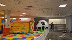Indoor play area with inflatable structures, colorful foam mat flooring, and a person organizing shelves in the background.