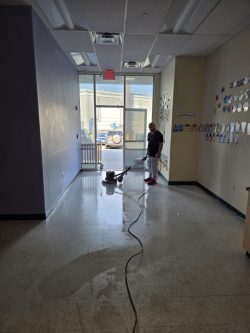 A person uses a floor buffer to clean the hallway of a building with sunlight coming through the glass doors.