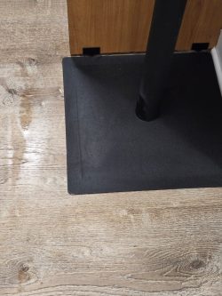 A black metal square base stands on a light wood floor next to a wooden and white wall corner.