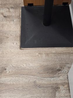 Square black metal table base on a light wood-patterned floor next to a wooden and white vertical surface.