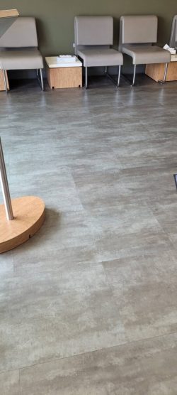 Gray chairs and wooden tables line a wall in a waiting area with gray tile flooring.