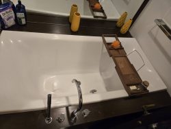 White bathtub with a wood bath tray, orange sponge, soap, and yellow vases on a dark countertop, beneath a large mirror.