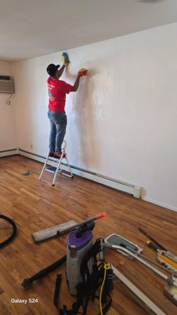 Person standing on a step stool cleans a white wall in a room with hardwood floors and cleaning tools on the floor.